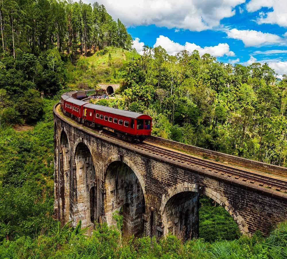 A red train crossing the Nine Arches Bridge in Ella, Sri Lanka, with the tunnel and jungle behind